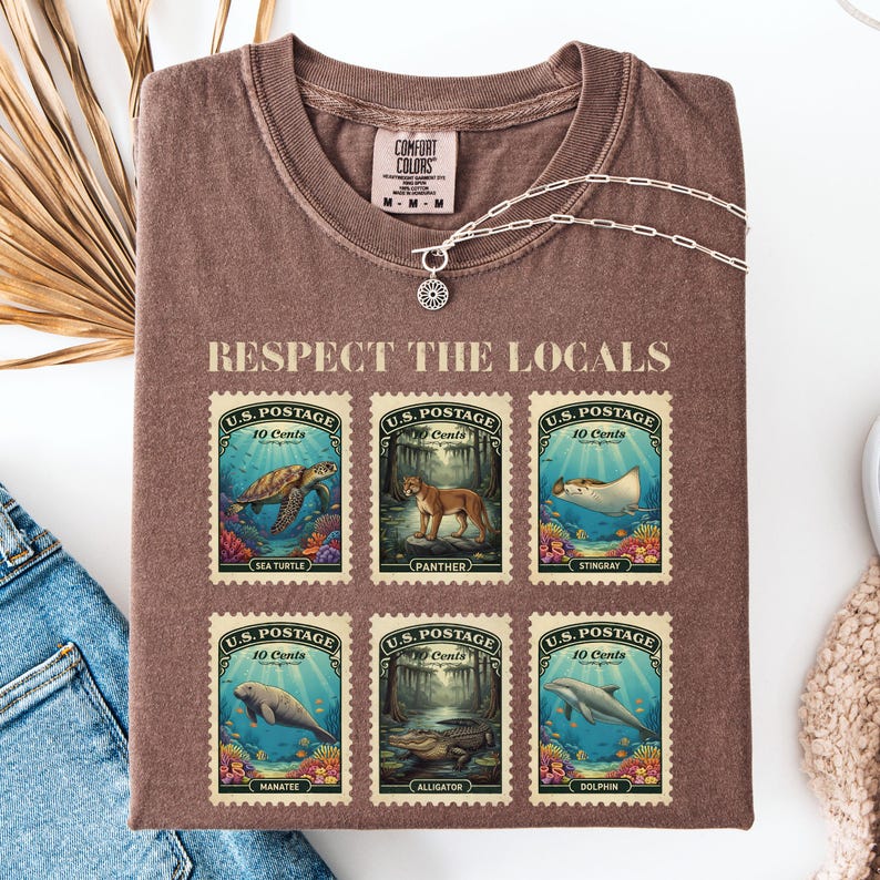 May include: Brown t-shirt with the text "RESPECT THE LOCALS" and six postage stamp designs featuring sea creatures and animals. The stamps read "U.S. Postage 10 Cents". A silver necklace with a pendant is on the shirt.