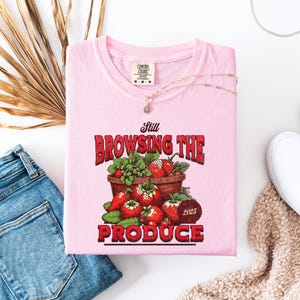 May include: A light pink t-shirt with the words "Still Browsing The Produce" in red lettering, featuring a graphic of strawberries in a pot. A gold necklace and a pair of blue jeans are next to the shirt.