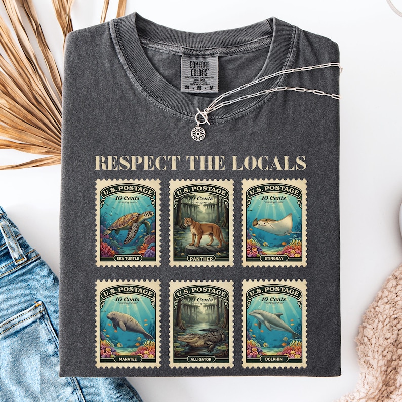 May include: Dark gray Comfort Colors t-shirt with the text "RESPECT THE LOCALS" above six postage stamp designs. The stamps feature illustrations of sea creatures and animals, including a sea turtle, panther, and dolphin.