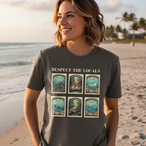 May include: Gray t-shirt with the text "RESPECT THE LOCALS" above six vintage-style postage stamps. The stamps feature illustrations of marine life and forest animals. The shirt is worn on a beach.