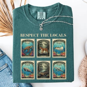 May include: Teal Comfort Colors t-shirt with the text "RESPECT THE LOCALS" and six postage stamp designs featuring sea creatures and animals. A silver necklace with a sun pendant is on top of the shirt.