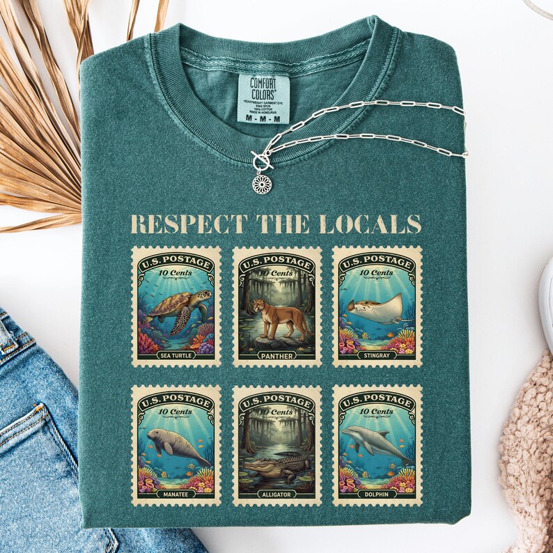 May include: Teal Comfort Colors t-shirt with the text "RESPECT THE LOCALS" and six postage stamp designs featuring sea creatures and animals. A silver necklace with a sun pendant is on top of the shirt.