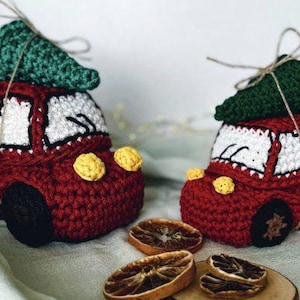 Könnte beinhalten: Zwei handgefertigte gehäkelte Spielzeugautos in Rot, Weiß und Schwarz, jedes mit einem grünen gehäkelten Weihnachtsbaum gekrönt. Die Autos haben gelbe Akzente und braune Räder. Getrocknete Orangenscheiben im Vordergrund. Eine festliche Weihnachtsdekoration.