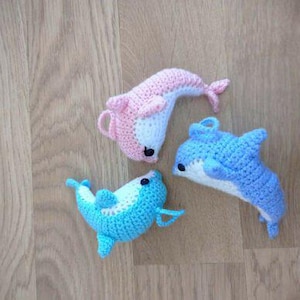 Puede incluir: Tres delfines de ganchillo hechos a mano en tonos rosa, azul y blanco están dispuestos sobre una superficie de madera. Los delfines tienen ojos negros y están elaborados con costuras detalladas, mostrando un tema acuático caprichoso.