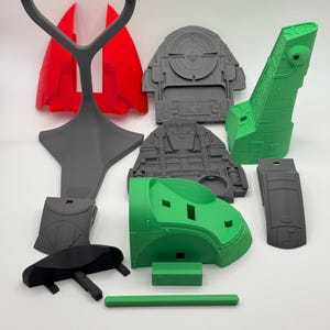 Puede incluir: Varias piezas de modelo impresas en 3D en rojo, gris, verde y negro. Los componentes, con formas geom&eacute;tricas y dise&ntilde;os detallados, parecen ser de un modelo m&aacute;s grande. Las piezas est&aacute;n dispuestas sobre una superficie blanca.