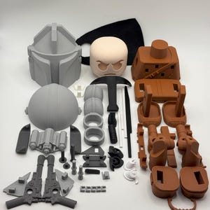 Puede incluir: Un conjunto de piezas sin montar, impresas en 3D, en varios colores, incluyendo gris, marr&oacute;n, negro y blanco. Los componentes, como un casco y armas, sugieren un tema Mandaloriano. Las piezas est&aacute;n dispuestas sobre una superficie blanca.