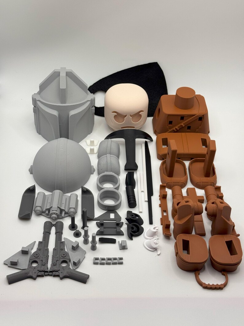Puede incluir: Un conjunto de piezas sin montar, impresas en 3D, en varios colores, incluyendo gris, marr&oacute;n, negro y blanco. Los componentes, como un casco y armas, sugieren un tema Mandaloriano. Las piezas est&aacute;n dispuestas sobre una superficie blanca.