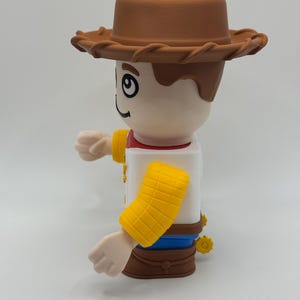 Op de afbeelding: Speelgoedfiguur van een cowboy-personage, met een bruine hoed, een wit shirt met een rode kraag en gele mouwmanchetten. De figuur heeft een lachende uitdrukking en de armen uitgestrekt.