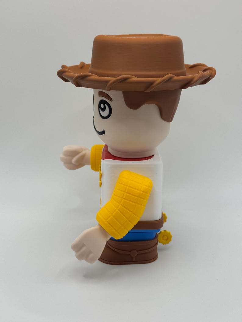 Op de afbeelding: Speelgoedfiguur van een cowboy-personage, met een bruine hoed, een wit shirt met een rode kraag en gele mouwmanchetten. De figuur heeft een lachende uitdrukking en de armen uitgestrekt.