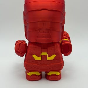 Puede incluir: Figura roja de superh&eacute;roe con detalles amarillos. La figura tiene un dise&ntilde;o en bloques, con casco y traje. Las costuras y texturas son visibles. El dise&ntilde;o sugiere un objeto de colecci&oacute;n o decorativo, posiblemente un personaje de c&oacute;mic.