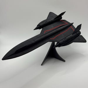 Puede incluir: Modelo negro de un avión SR-71 Blackbird con líneas rojas. El modelo está montado sobre un soporte negro. El avión tiene un cuerpo largo y delgado con alas y aletas de cola. Réplica detallada del icónico avión de reconocimiento.