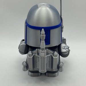 Peut inclure: Figurine argent&eacute;e et bleue d'un personnage avec un casque et un jetpack. La figurine a une t&ecirc;te arrondie avec une bande bleue et une antenne bleue. Le jetpack et d'autres d&eacute;tails sont &eacute;galement argent&eacute;s et bleus.