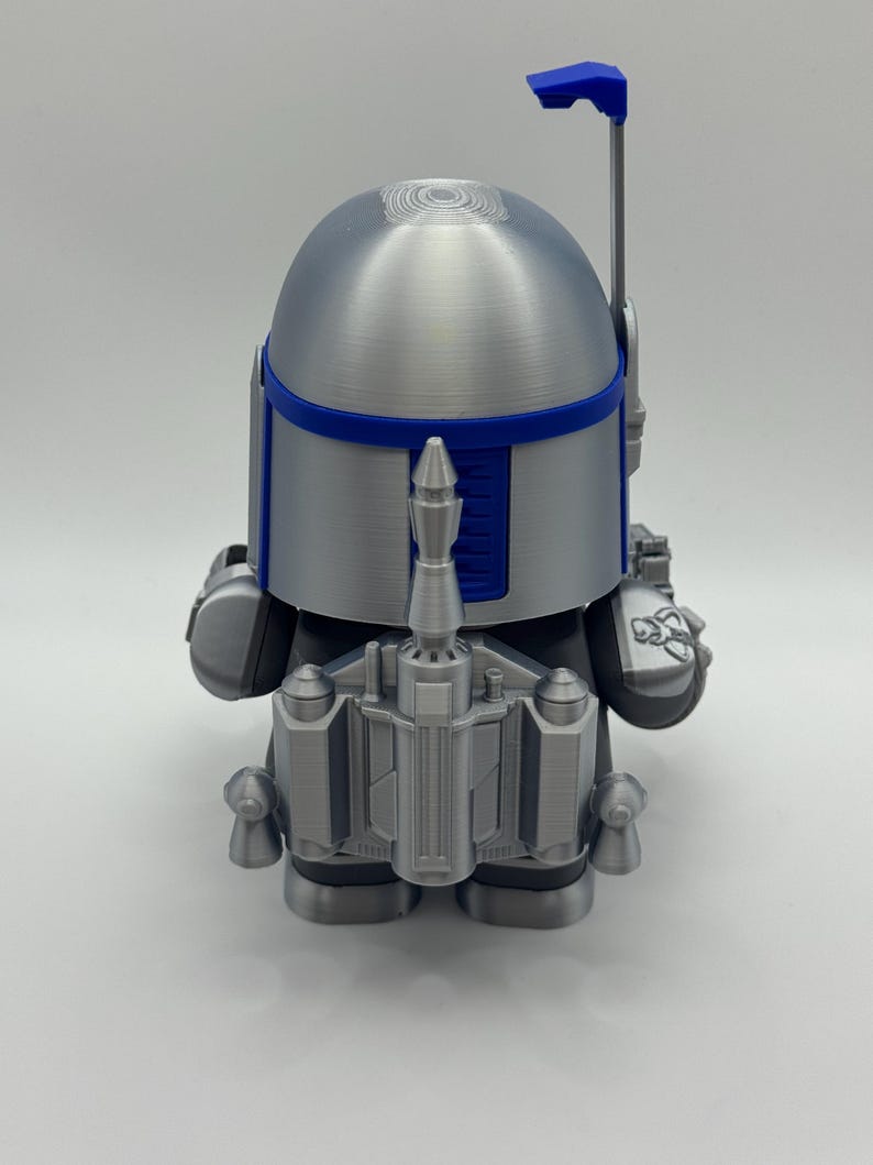 Peut inclure: Figurine argent&eacute;e et bleue d'un personnage avec un casque et un jetpack. La figurine a une t&ecirc;te arrondie avec une bande bleue et une antenne bleue. Le jetpack et d'autres d&eacute;tails sont &eacute;galement argent&eacute;s et bleus.