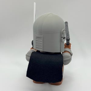 May include: A grey and brown action figure of a character wearing a helmet and a black cape. The figure is holding a weapon in its right hand.