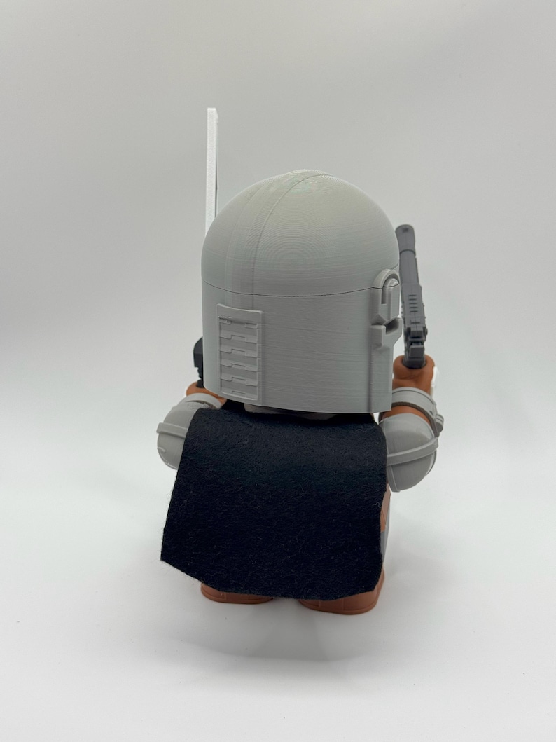 May include: A grey and brown action figure of a character wearing a helmet and a black cape. The figure is holding a weapon in its right hand.