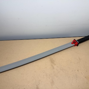May include: A long sword with a smooth gray blade, a bright red tsuba guard featuring a black circular detail, and a textured black handle. The sword is positioned diagonally across the frame, showcasing its full length and distinct color segments.