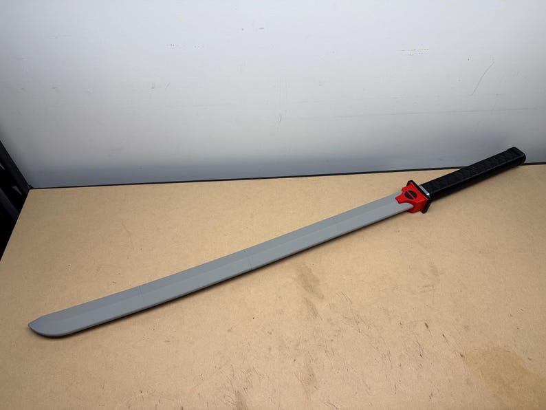 May include: A long sword with a smooth gray blade, a bright red tsuba guard featuring a black circular detail, and a textured black handle. The sword is positioned diagonally across the frame, showcasing its full length and distinct color segments.