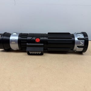 Puede incluir: Un accesorio de sable de luz negro y plateado con un bot&oacute;n rojo y un agarre rectangular. El sable de luz tiene un cuerpo cil&iacute;ndrico con detalles plateados en ambos extremos y un emisor negro.