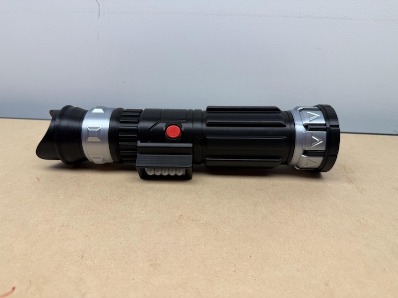 Puede incluir: Un accesorio de sable de luz negro y plateado con un bot&oacute;n rojo y un agarre rectangular. El sable de luz tiene un cuerpo cil&iacute;ndrico con detalles plateados en ambos extremos y un emisor negro.