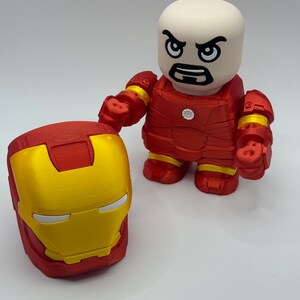 Puede incluir: Una figura y un casco de Iron Man impresos en 3D. La figura es roja con detalles amarillos y una cabeza blanca con una expresi&oacute;n severa. El casco es rojo y dorado. Los art&iacute;culos est&aacute;n sobre una superficie blanca.
