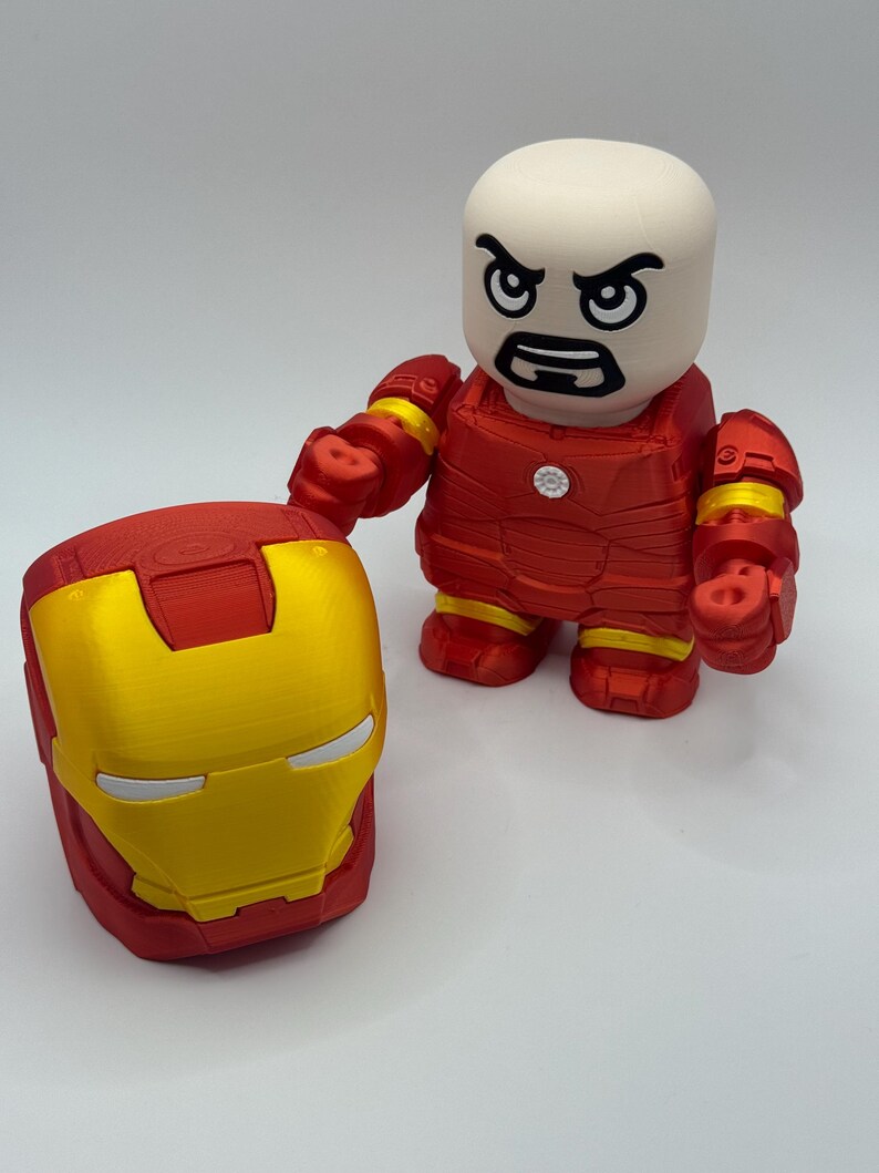 Puede incluir: Una figura y un casco de Iron Man impresos en 3D. La figura es roja con detalles amarillos y una cabeza blanca con una expresi&oacute;n severa. El casco es rojo y dorado. Los art&iacute;culos est&aacute;n sobre una superficie blanca.