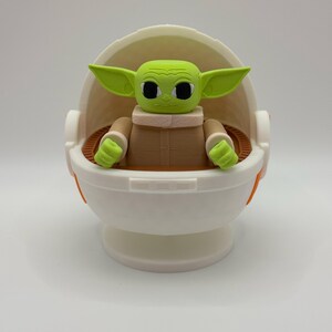 May include: A green and brown figurine of a character from the Star Wars franchise, sitting in a white and orange egg-shaped pod. The character has large eyes and a small mouth.