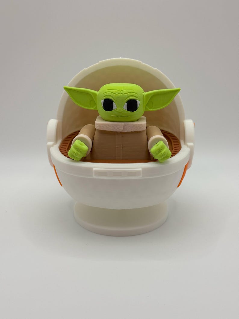 May include: A green and brown figurine of a character from the Star Wars franchise, sitting in a white and orange egg-shaped pod. The character has large eyes and a small mouth.