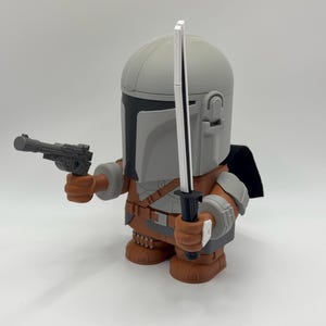 Puede incluir: Figura impresa en 3D de un personaje con casco y armadura, sosteniendo una pistola bl&aacute;ster y una espada. La figura es principalmente gris, marr&oacute;n y negra, con una capa negra. La figura est&aacute; sobre una superficie blanca.
