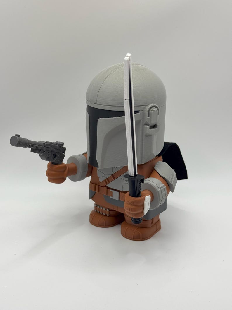 Puede incluir: Figura impresa en 3D de un personaje con casco y armadura, sosteniendo una pistola bl&aacute;ster y una espada. La figura es principalmente gris, marr&oacute;n y negra, con una capa negra. La figura est&aacute; sobre una superficie blanca.