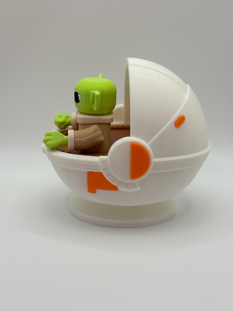 May include: A green and brown Yoda character figurine sitting in a white and orange spaceship. The spaceship has a dome-shaped top and a round base.