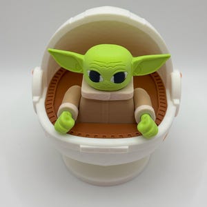 May include: A green and brown toy figure of a character from the Star Wars franchise, sitting in a white egg-shaped pod. The figure has large eyes and a small mouth.