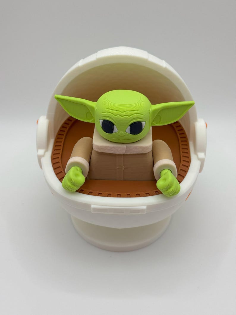 May include: A green and brown toy figure of a character from the Star Wars franchise, sitting in a white egg-shaped pod. The figure has large eyes and a small mouth.