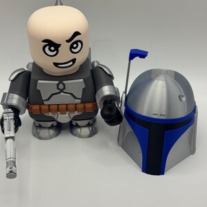 Peut inclure: Une figurine stylis&eacute;e et un casque, rappelant un personnage de Star Wars. La figurine est principalement grise et noire, avec une t&ecirc;te beige et un blaster. Le casque est argent&eacute; avec des touches de bleu.
