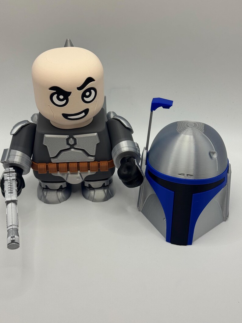 Peut inclure: Une figurine stylis&eacute;e et un casque, rappelant un personnage de Star Wars. La figurine est principalement grise et noire, avec une t&ecirc;te beige et un blaster. Le casque est argent&eacute; avec des touches de bleu.