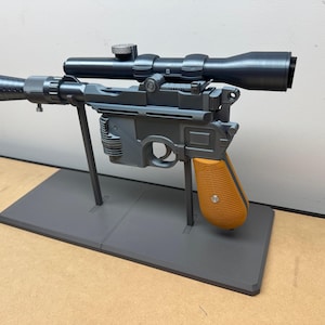May include: A grey and black replica blaster pistol with a scope and orange grip. The pistol is mounted on a grey stand. The barrel has an orange tip.
