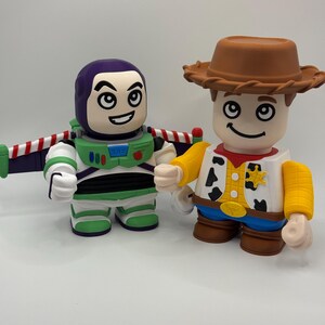 Op de afbeelding: Twee gestileerde beeldjes van Toy Story-personages Buzz Lightyear en Woody. Buzz draagt een groen en wit ruimtepak met paarse accenten en vleugels. Woody draagt een cowboyhoed, vest en een blauwe spijkerbroek. Beide figuren hebben lachende gezichten.