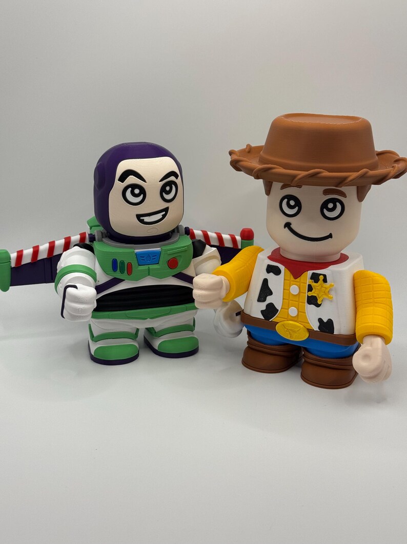 Op de afbeelding: Twee gestileerde beeldjes van Toy Story-personages Buzz Lightyear en Woody. Buzz draagt een groen en wit ruimtepak met paarse accenten en vleugels. Woody draagt een cowboyhoed, vest en een blauwe spijkerbroek. Beide figuren hebben lachende gezichten.