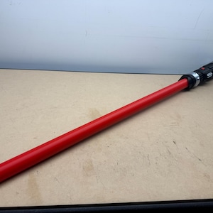 Puede incluir: Un sable de luz rojo con una empu&ntilde;adura negra y plateada. El sable de luz est&aacute; sobre una superficie marr&oacute;n claro. El sable de luz tiene forma cil&iacute;ndrica y larga, con una punta redondeada.
