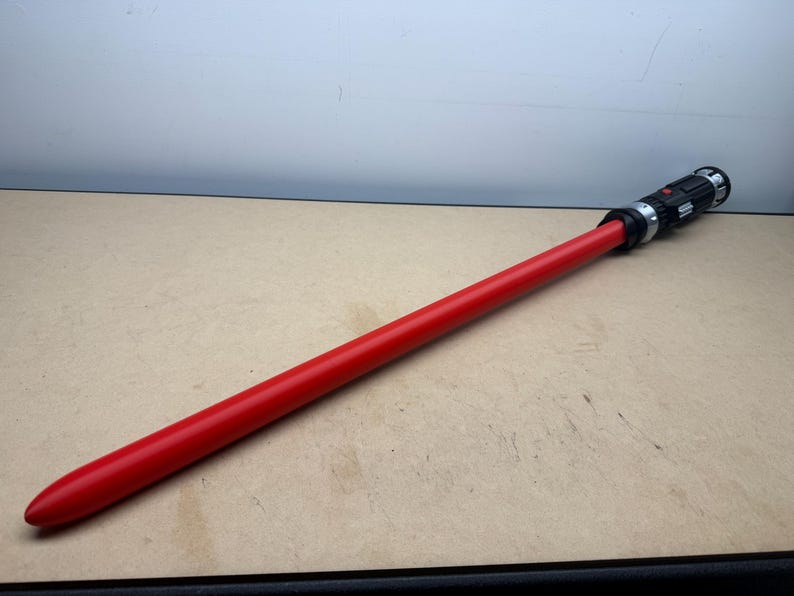 Puede incluir: Un sable de luz rojo con una empu&ntilde;adura negra y plateada. El sable de luz est&aacute; sobre una superficie marr&oacute;n claro. El sable de luz tiene forma cil&iacute;ndrica y larga, con una punta redondeada.