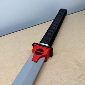 May include: A sword with a long, light gray blade and a black handle featuring a textured, crisscross pattern. The guard is bright red with a circular black accent in the center, connecting the handle to the blade in a straight, linear design.