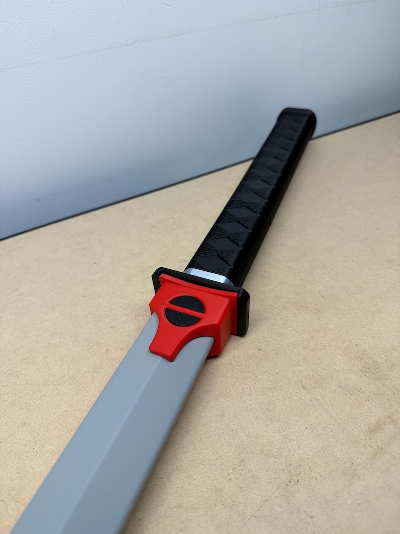 May include: A sword with a long, light gray blade and a black handle featuring a textured, crisscross pattern. The guard is bright red with a circular black accent in the center, connecting the handle to the blade in a straight, linear design.