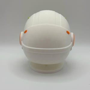 May include: White astronaut figurine with orange accents and green details on the top of the head. The figurine is standing on a white base.