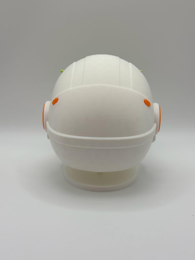 May include: White astronaut figurine with orange accents and green details on the top of the head. The figurine is standing on a white base.