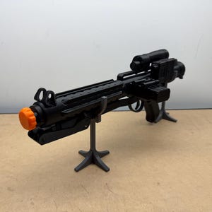 May include: Black toy blaster rifle with an orange tip, mounted on a black stand. The rifle has a scope and various attachments. The background is a plain white wall and a beige surface.
