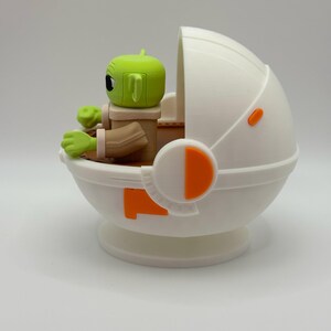 Puede incluir: Una figura de personaje de piel verde con orejas grandes, sentada en un portabeb&eacute;s blanco y naranja en forma de c&aacute;psula. La figura tiene ojos negros grandes y viste un atuendo marr&oacute;n. El portabeb&eacute;s tiene detalles en naranja y un dise&ntilde;o redondeado.