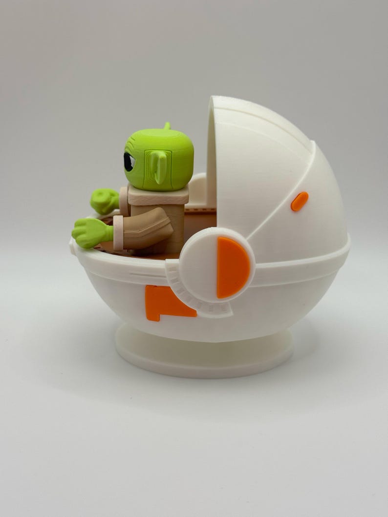 Puede incluir: Una figura de personaje de piel verde con orejas grandes, sentada en un portabeb&eacute;s blanco y naranja en forma de c&aacute;psula. La figura tiene ojos negros grandes y viste un atuendo marr&oacute;n. El portabeb&eacute;s tiene detalles en naranja y un dise&ntilde;o redondeado.