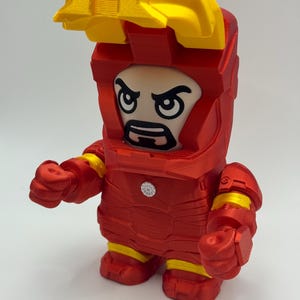Puede incluir: Una figura de Iron Man roja y amarilla con un dise&ntilde;o en bloques. La figura tiene un casco amarillo y una armadura roja con detalles amarillos. La cara es una versi&oacute;n de dibujos animados de la cara de Iron Man.