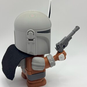 May include: A grey and brown action figure of a Mandalorian with a black cape and a grey blaster pistol. The figure is holding the blaster in its right hand.