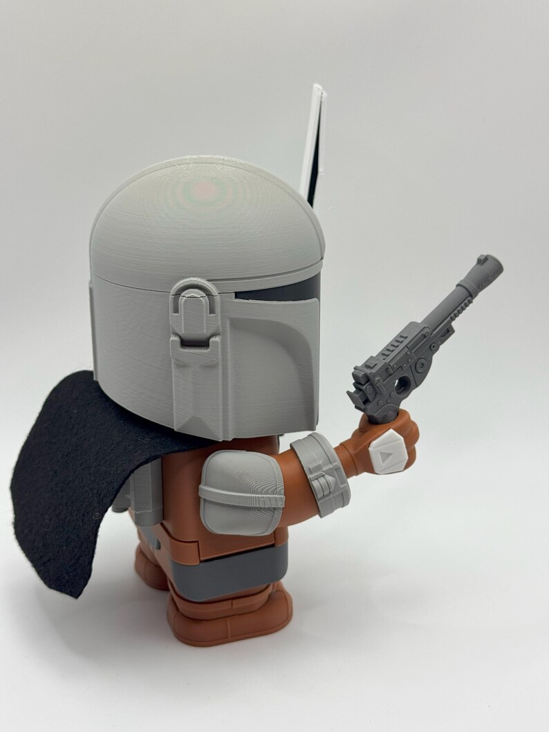 May include: A grey and brown action figure of a Mandalorian with a black cape and a grey blaster pistol. The figure is holding the blaster in its right hand.