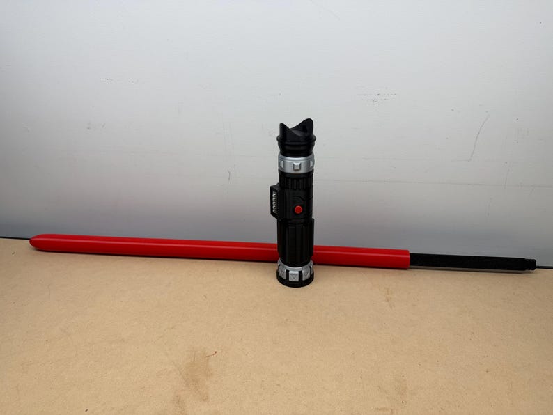 Puede incluir: Un sable de luz de juguete negro y plateado con una hoja roja. El mango presenta detalles plateados y un bot&oacute;n rojo. La hoja roja est&aacute; horizontal, con el mango vertical.